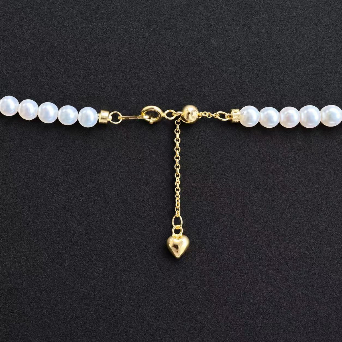 Close-up of the YUKI Baby freshwater pearl necklace’s gold-plated clasp, sliding adjustment chain and heart-shaped charm on a black background.