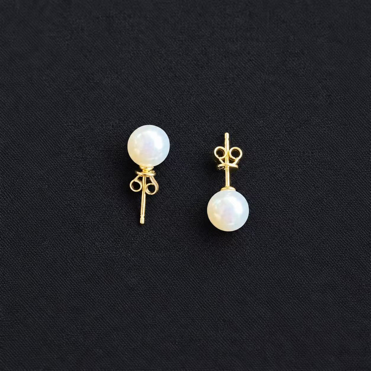 Pair of white freshwater pearl stud earrings showing gold-tone posts and butterfly backs, displayed on a black fabric background.