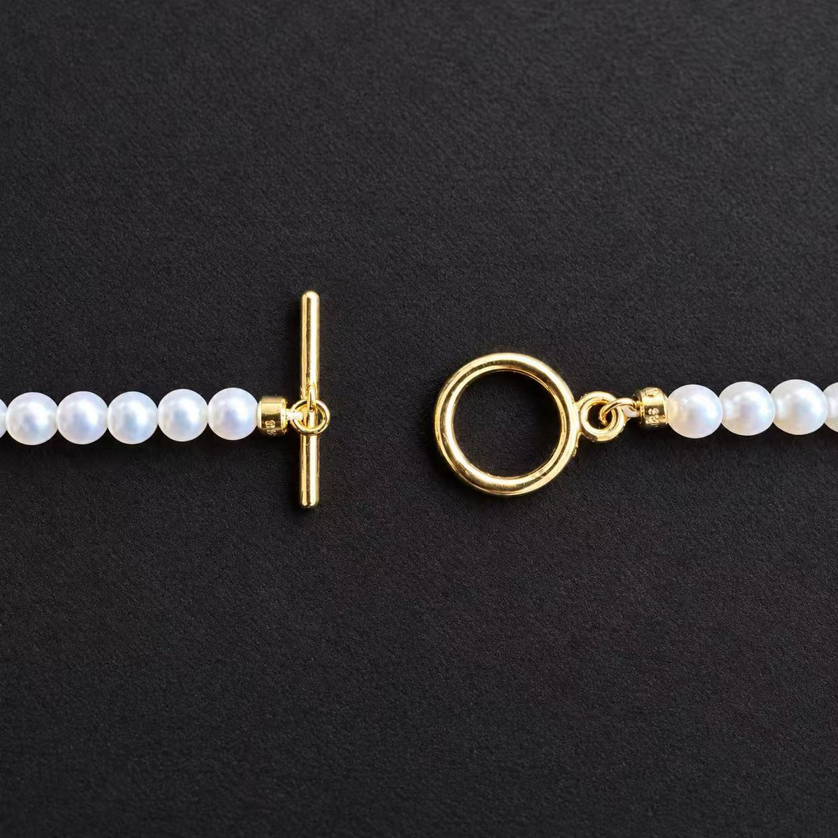 Close-up of the Baby freshwater pearl necklace’s gold-tone toggle clasp, showing the separate bar and ring on a black background.