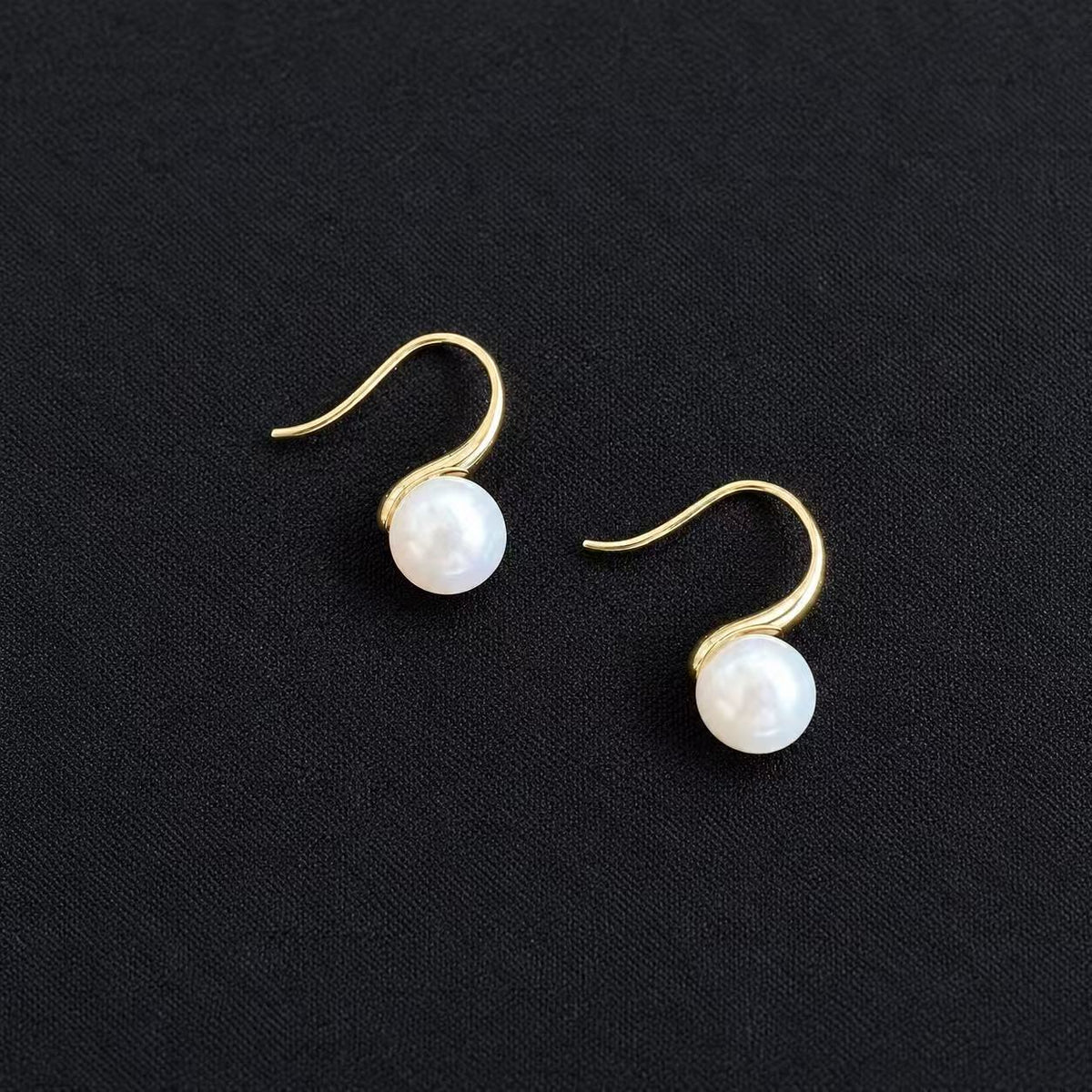 Pair of YUKI Arc earrings with white freshwater pearls and curved gold-plated silver hooks, displayed on a black background.