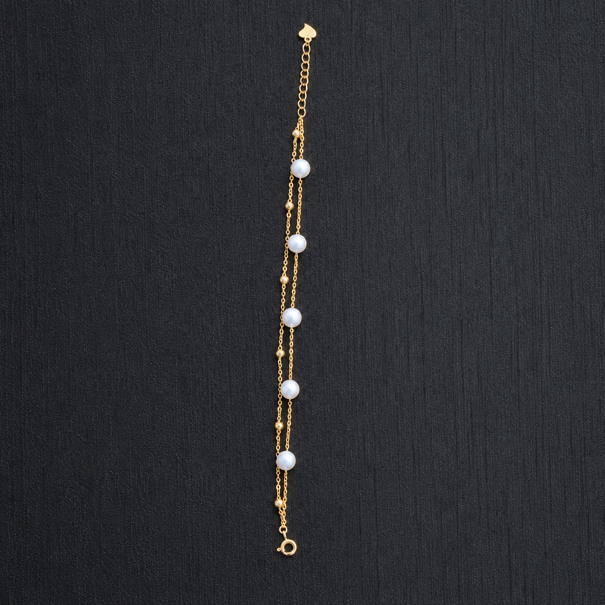 YUKI Double Chain bracelet with white freshwater pearls and small gold-tone beads, laid flat on a dark textured background, showing the clasp and extender chain.