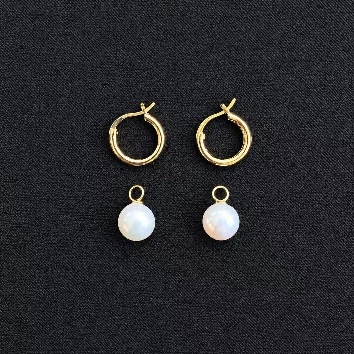 Pair of gold-tone hoop earrings with two detachable white freshwater pearl charms shown separately below the hoops on a black fabric background.