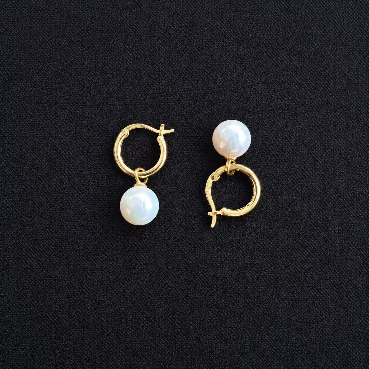 Pair of gold-tone hoop earrings with white freshwater pearl drops, displayed on a black fabric background.